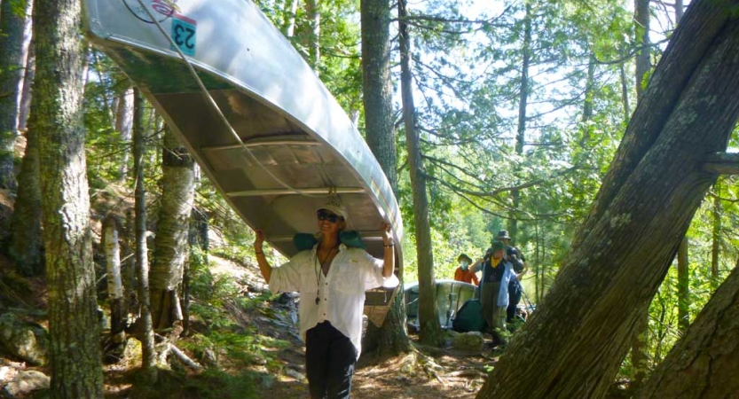 A person carries a canoe on their shoulders through a wooded area. 